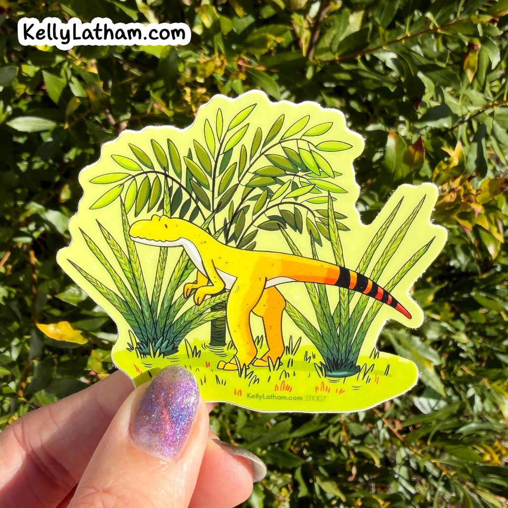 A hand with glittery purple nail polish holds a die-cut sticker featuring a yellow cartoon dinosaur among green plants. The sticker is photographed outdoors in front of dense green foliage, with the brand name KellyLatham.