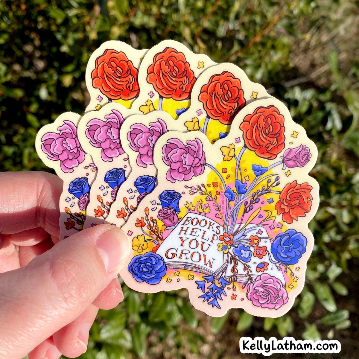 A hand holds a fanned-out set of die-cut stickers featuring a colorful floral and book illustration with the text 'BOOKS HELP YOU GROW'. The background is a blurred outdoor green garden, and a watermark 'KellyLatham.