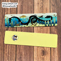 Two rectangular printed cards or bookmarks laid flat on a wooden surface. The top card shows an illustrated underwater scene with a black serpent-like creature, blue water, fish, and small marine details.
