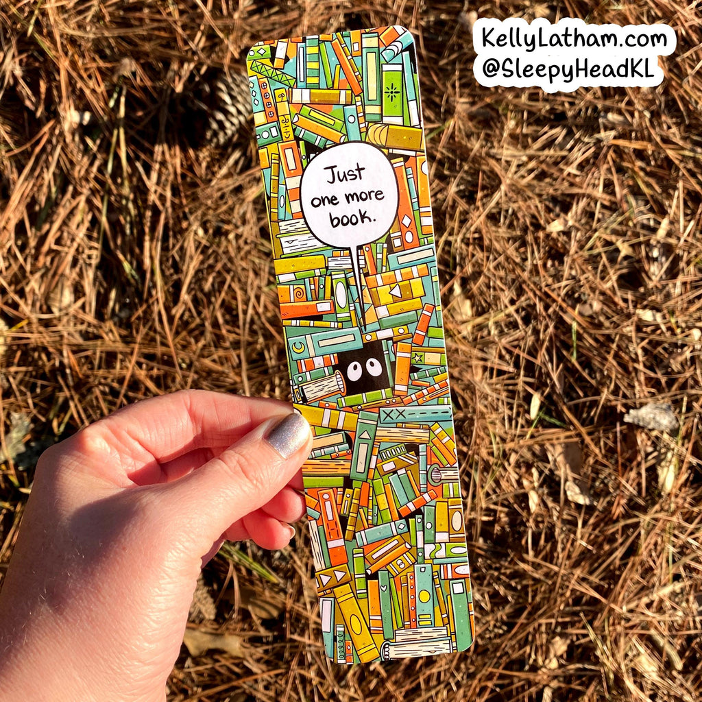 A hand holds a long, narrow bookmark or card featuring a colorful illustrated pattern of stacked books, with a speech bubble that says 'Just one more book.' The background is dry pine needles outdoors, and the text 'KellyLatham.