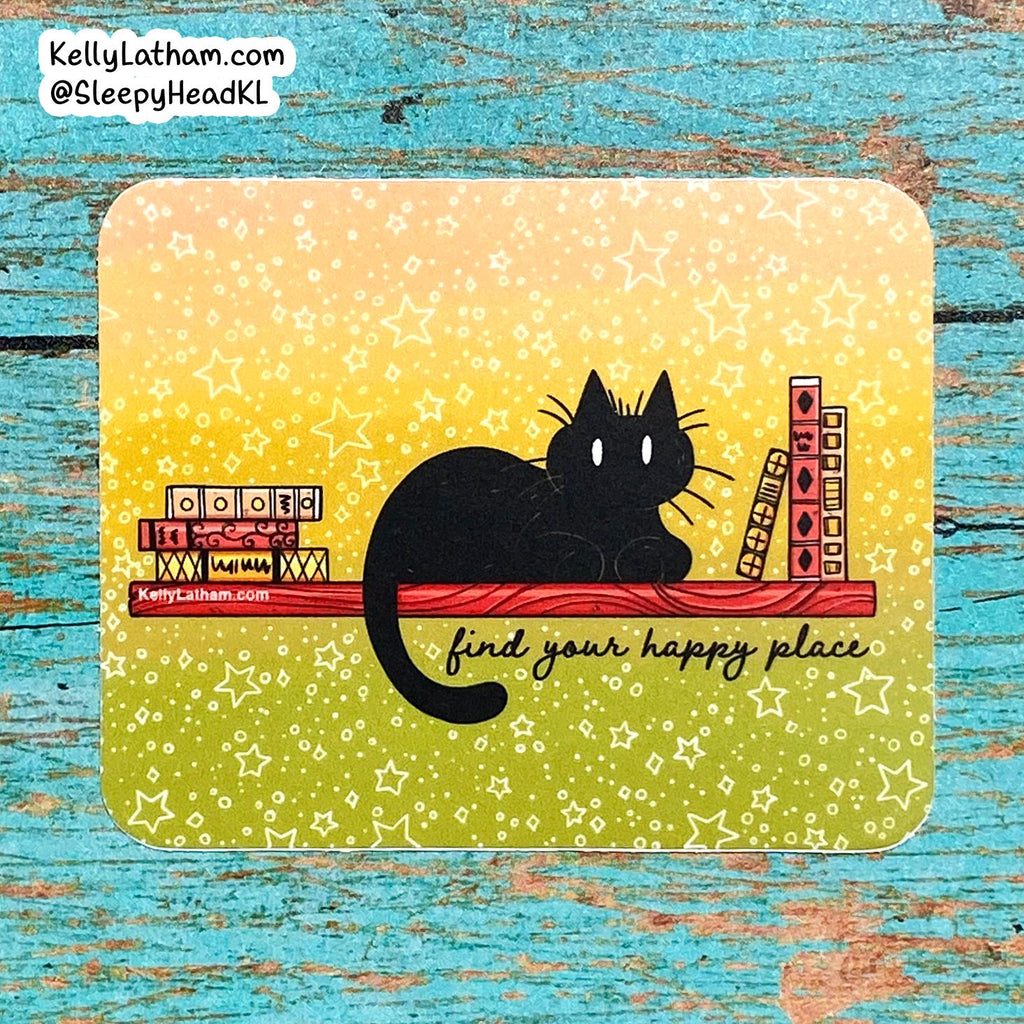 A square printed card or art piece with rounded corners, featuring a black cat sitting on a red shelf against a yellow-to-green gradient background with white stars. The card includes the phrase 'find your happy place' and is photographed on a distressed turquoise wooden surface.