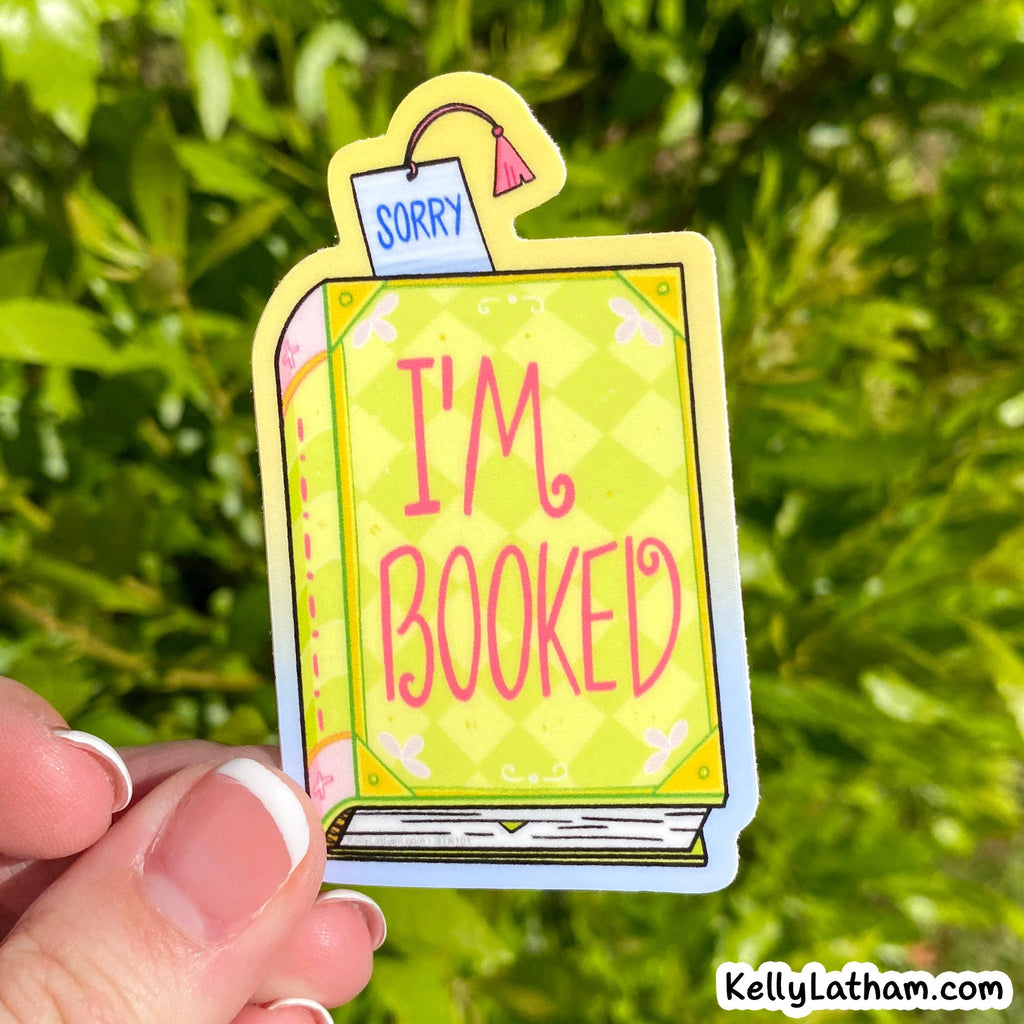 A hand holds a die-cut sticker shaped like a book with the words “I'M BOOKED” in pink on a yellow patterned cover. The sticker has a small “SORRY” note at the top and is photographed outdoors against a blurred green leafy background.