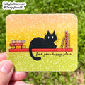 A hand holds a rectangular printed card or sticker outdoors against a blurred green grass background. The card has rounded corners and features a black cat sitting on a red shelf with books and the phrase 'find your happy place' on a yellow-to-green gradient background with white star doodles.