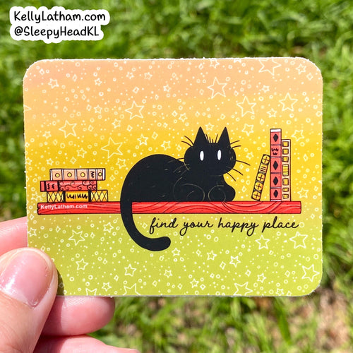 A hand holds a rectangular printed card or sticker outdoors against a blurred green grass background. The card has rounded corners and features a black cat sitting on a red shelf with books and the phrase 'find your happy place' on a yellow-to-green gradient background with white star doodles.