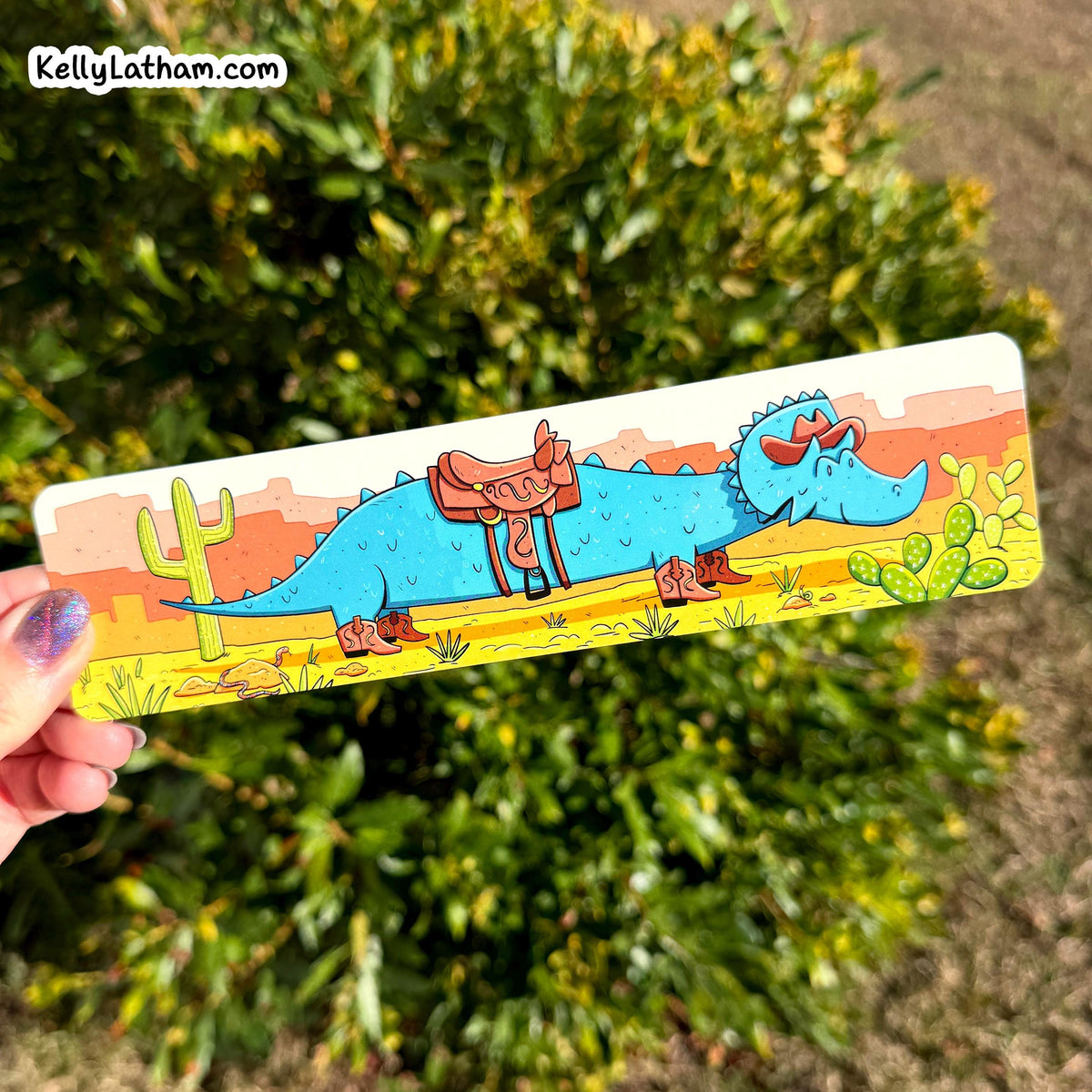 A hand holds a rectangular bookmark featuring a colorful cartoon illustration of a blue dinosaur with a brown saddle, boots, cactus, and desert landscape, set against a blurred green garden background.