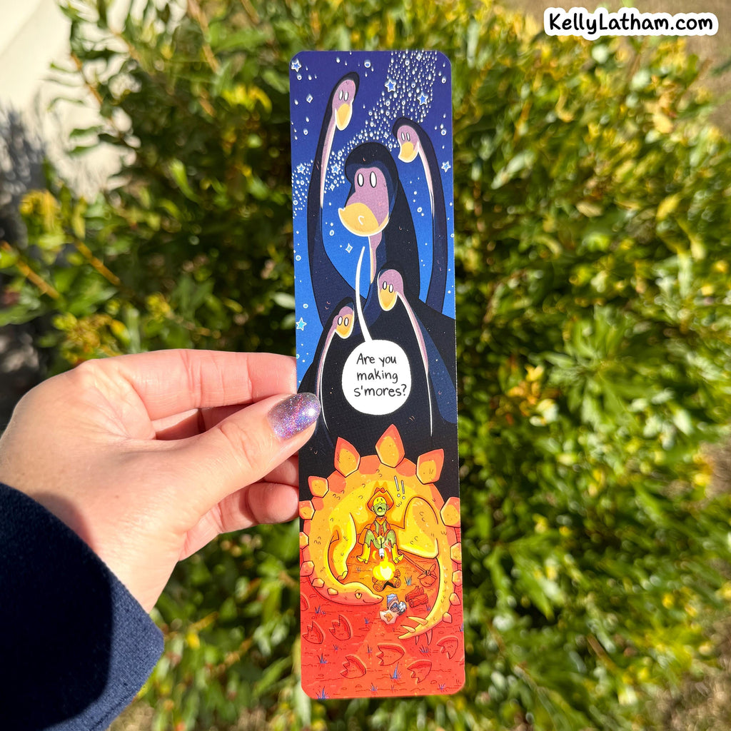 A hand holds a long, narrow illustrated card or bookmark outdoors in front of green foliage. The artwork shows a dark blue upper section with a stylized face and glowing moon-like circles, a white speech bubble reading 'Are you making s'mores?', and a fiery orange-red lower section with a meditating figure.