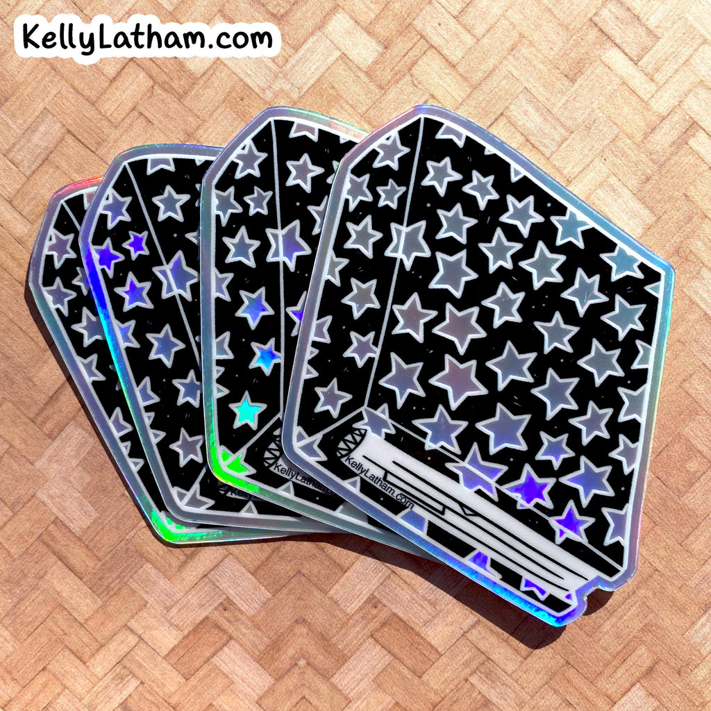 Four holographic credit card-sized stickers with a black background and silver star pattern, arranged in a fan on a light wood herringbone surface. The brand name KellyLatham.