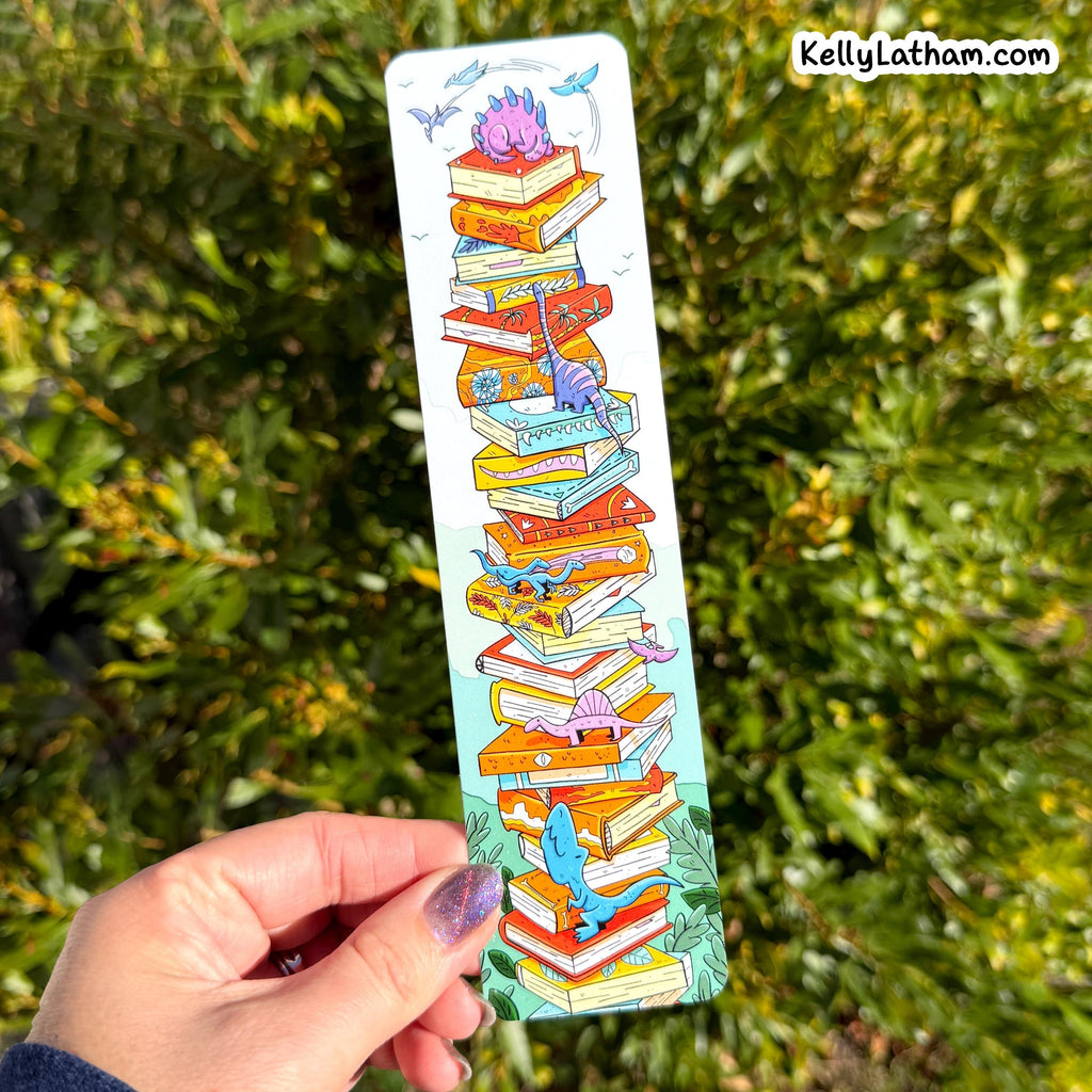 A hand holds a long, narrow illustrated bookmark or card featuring a stack of colorful books with small fantasy creatures, set against a blurred green outdoor background. The text 'KellyLatham.