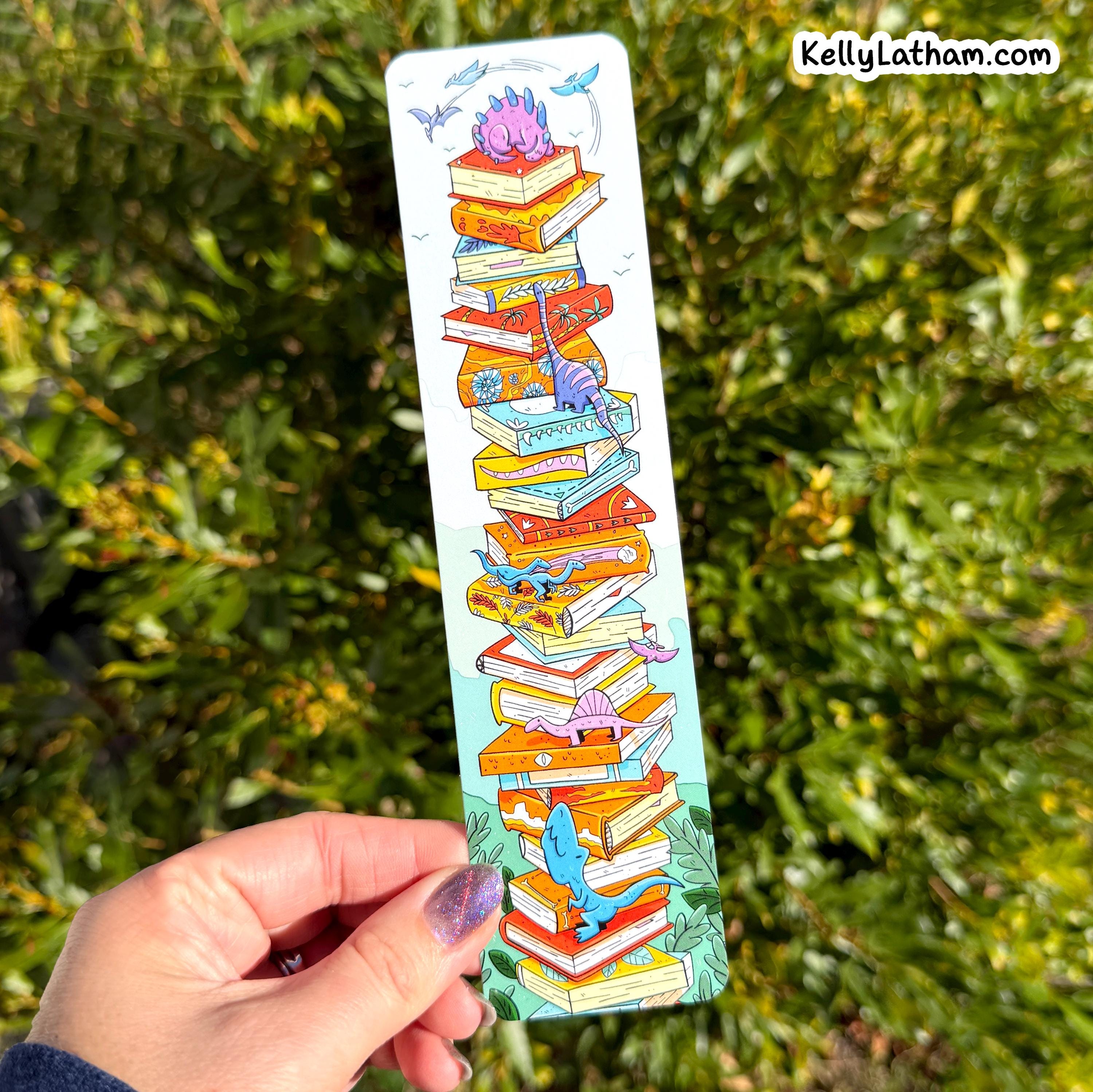 A hand holds a long, narrow illustrated bookmark or card featuring a stack of colorful books with small fantasy creatures, set against a blurred green outdoor background. The text 'KellyLatham.