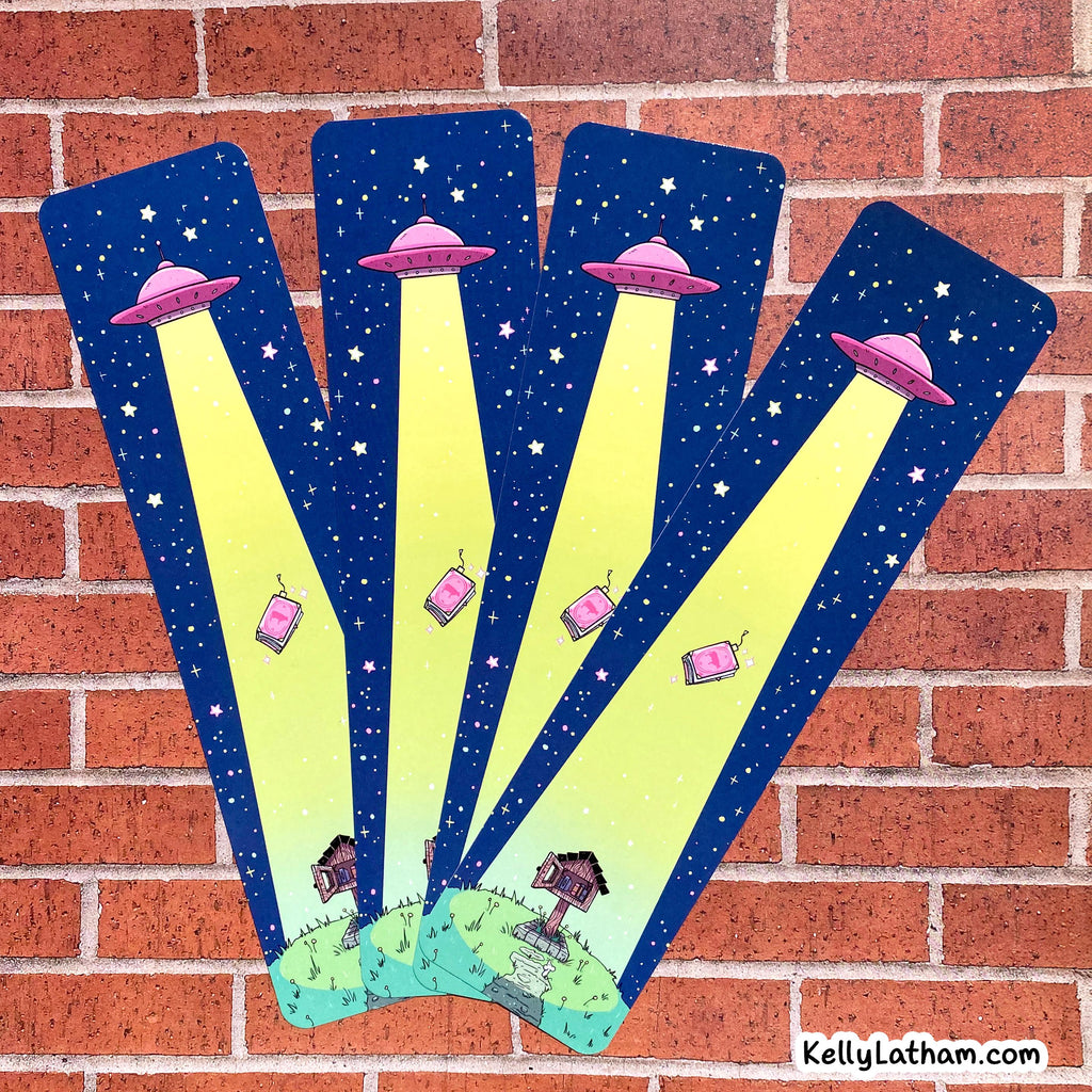 Four matching decorative wall decals or magnets featuring a whimsical space theme, fanned out against a red brick wall. The illustrations show pink flying saucers casting bright yellow spotlights over a starry dark blue night sky, with small houses on a grassy hill at the bottom.