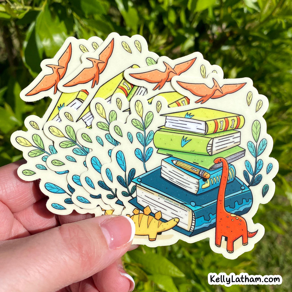 A hand holds a die-cut sticker featuring an illustrated stack of books, a small dinosaur, flying birds, and leafy vines. The background is a blurred green outdoor setting.