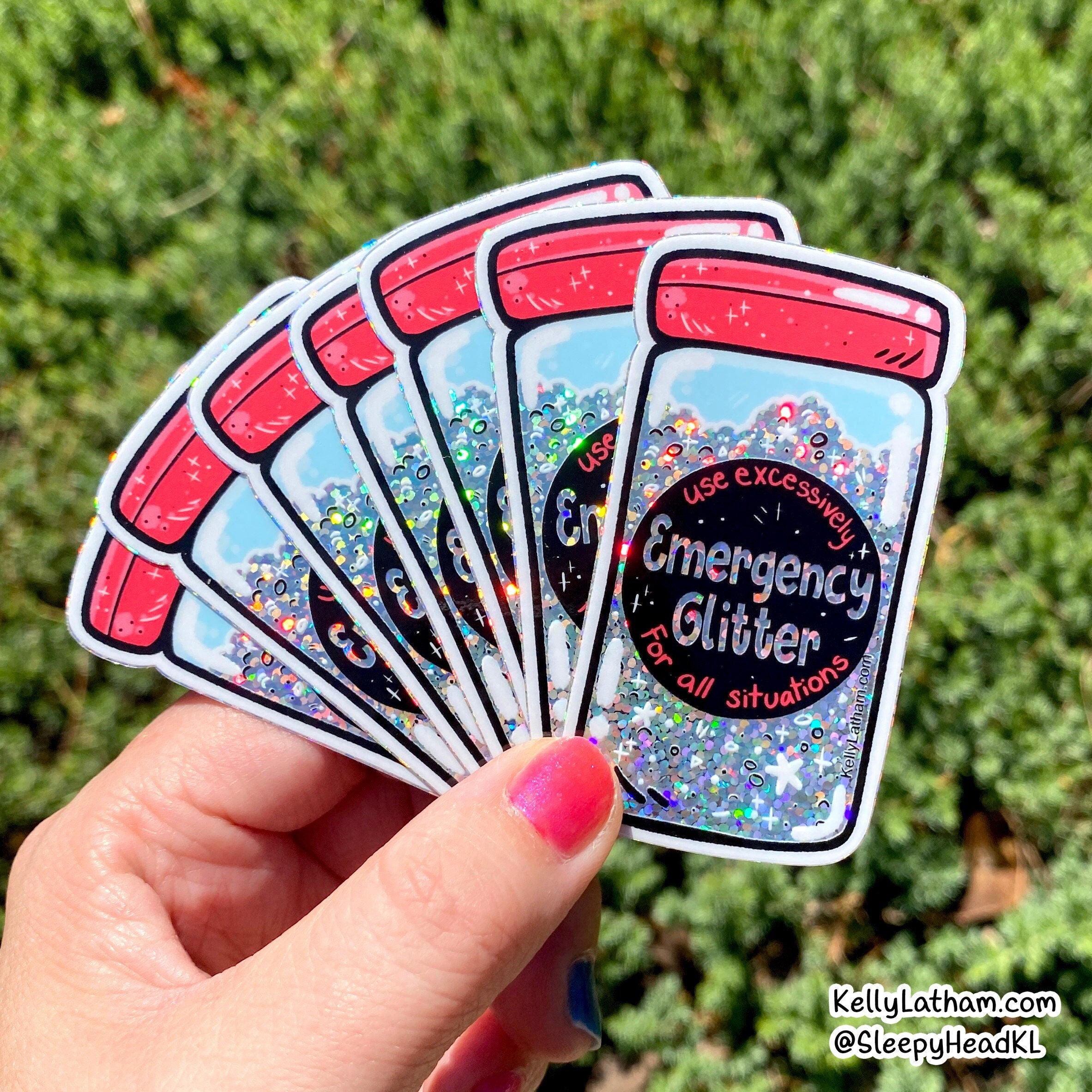 A hand holds a fan of five die-cut stickers shaped like glittery emergency supply jars, with the center sticker reading 'Emergency Glitter for all situations.' The background is blurred green foliage.