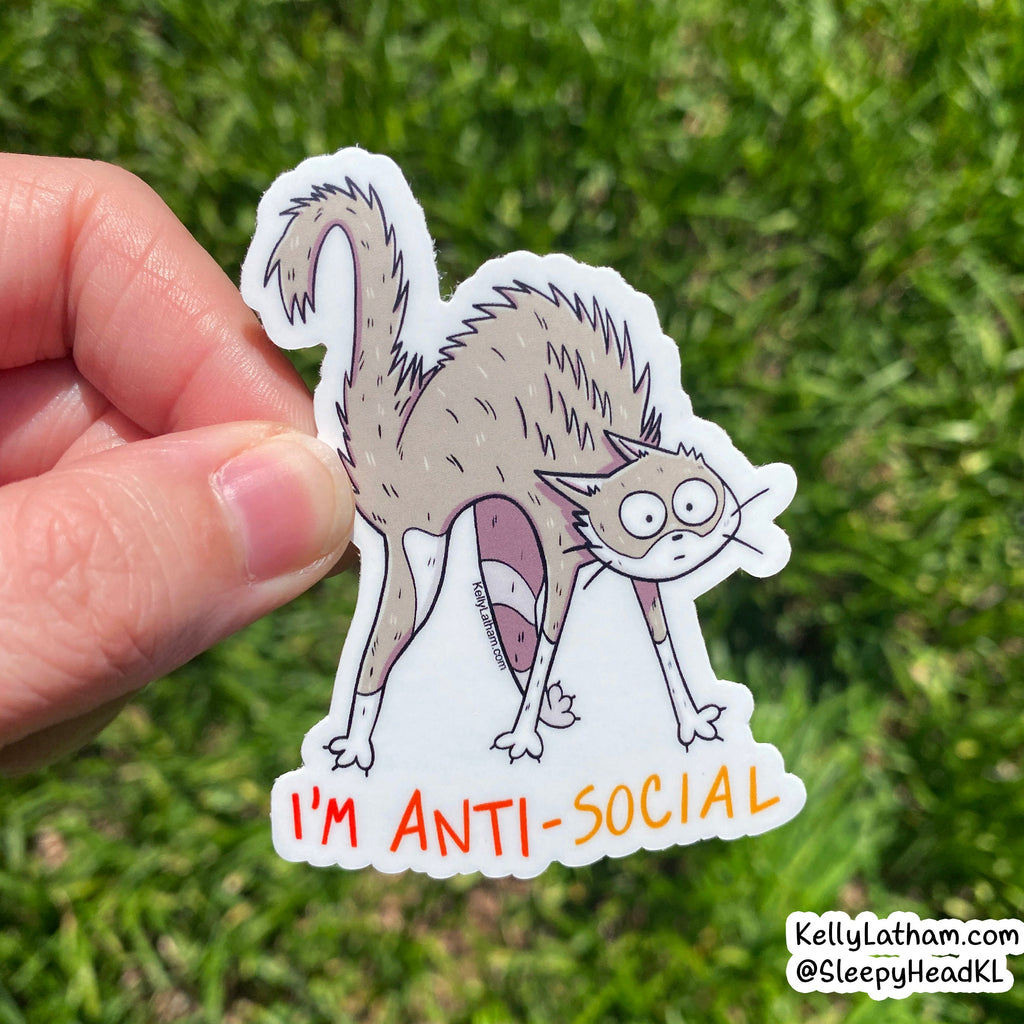 Cartoon cat sticker labeled 'I'm Anti-Social' held by a hand against a grass background