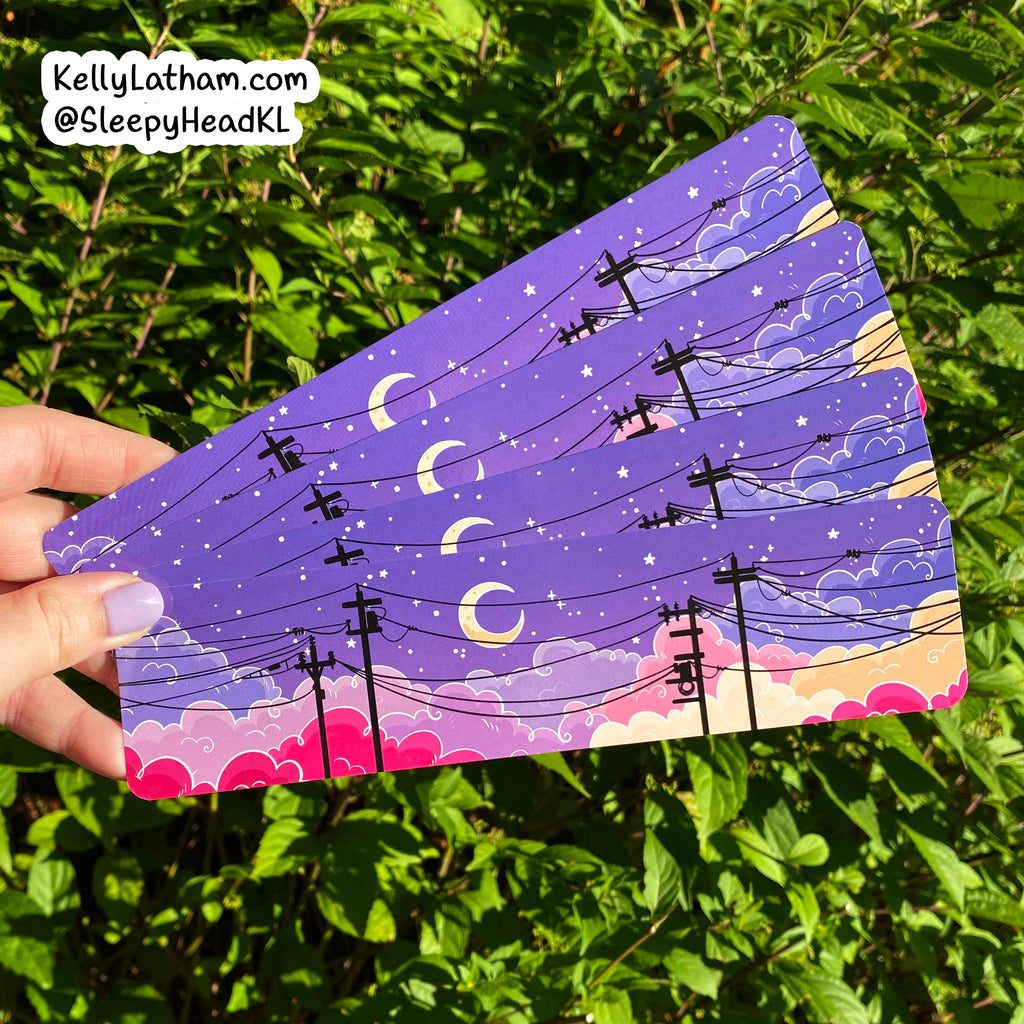 A hand holds a fan of rectangular art cards or stickers featuring a purple night sky with crescent moons, stars, clouds, and black utility poles, against a background of green leaves. A brand watermark in the top left reads KellyLatham.