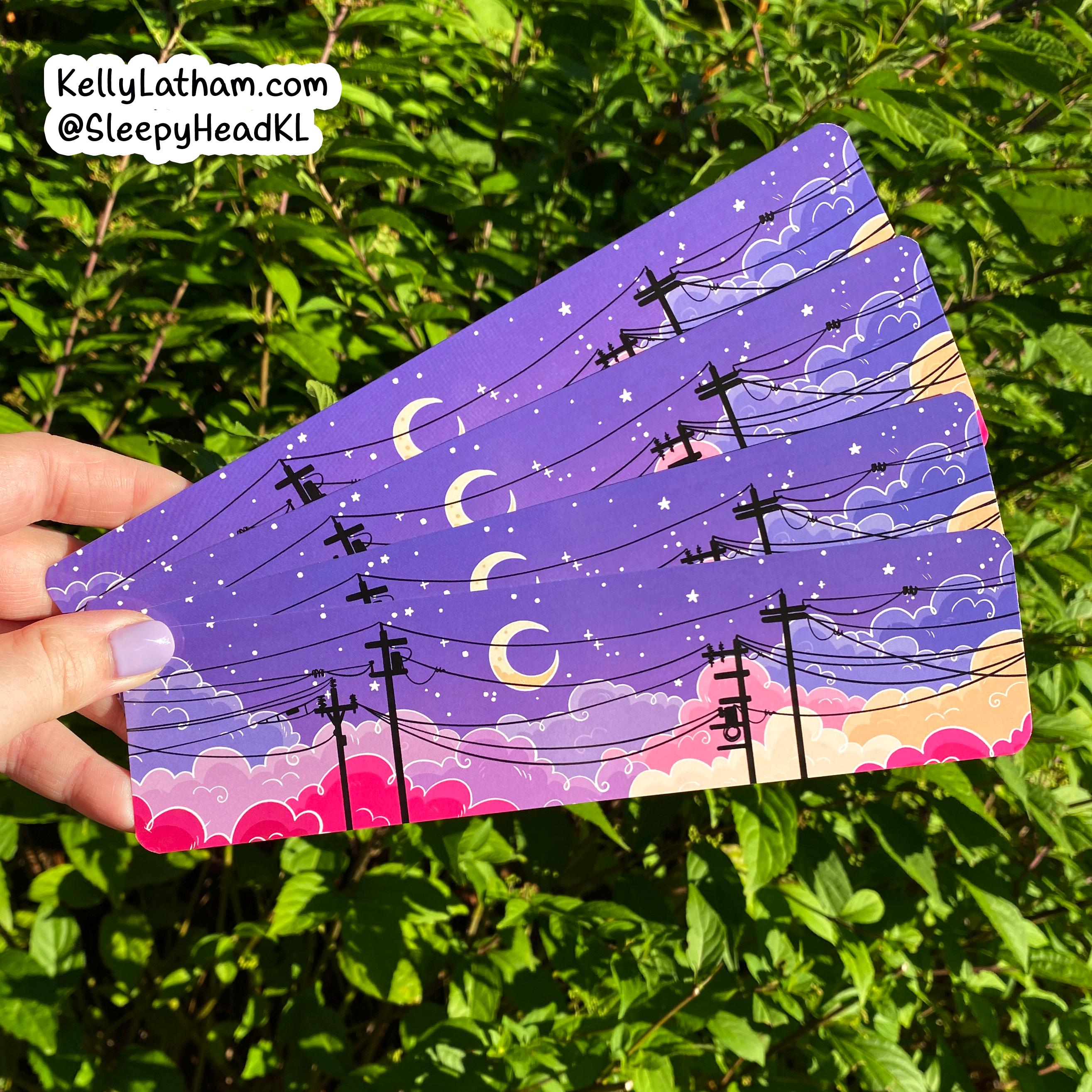 A hand holds a fan of rectangular art cards or stickers featuring a purple night sky with crescent moons, stars, clouds, and black utility poles, against a background of green leaves. A brand watermark in the top left reads KellyLatham.