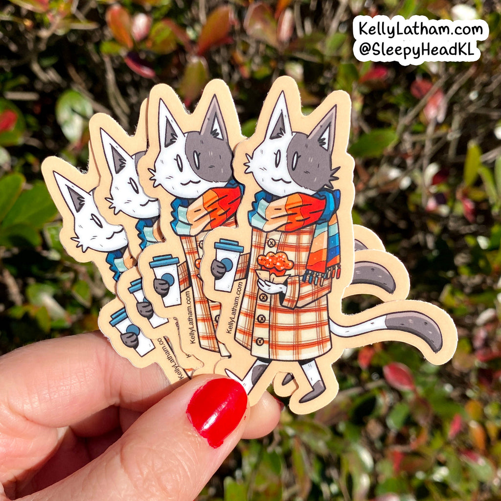 A hand with red nail polish holds four die-cut vinyl stickers featuring a cartoon cat in a plaid coat and colorful scarf, with a blurred outdoor garden background. A brand watermark in the top right reads KellyLatham.