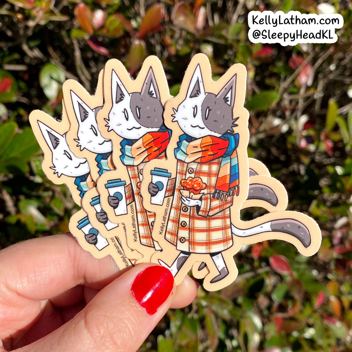 A hand with red nail polish holds four die-cut vinyl stickers featuring a cartoon cat in a plaid coat and colorful scarf, with a blurred outdoor garden background. A brand watermark in the top right reads KellyLatham.