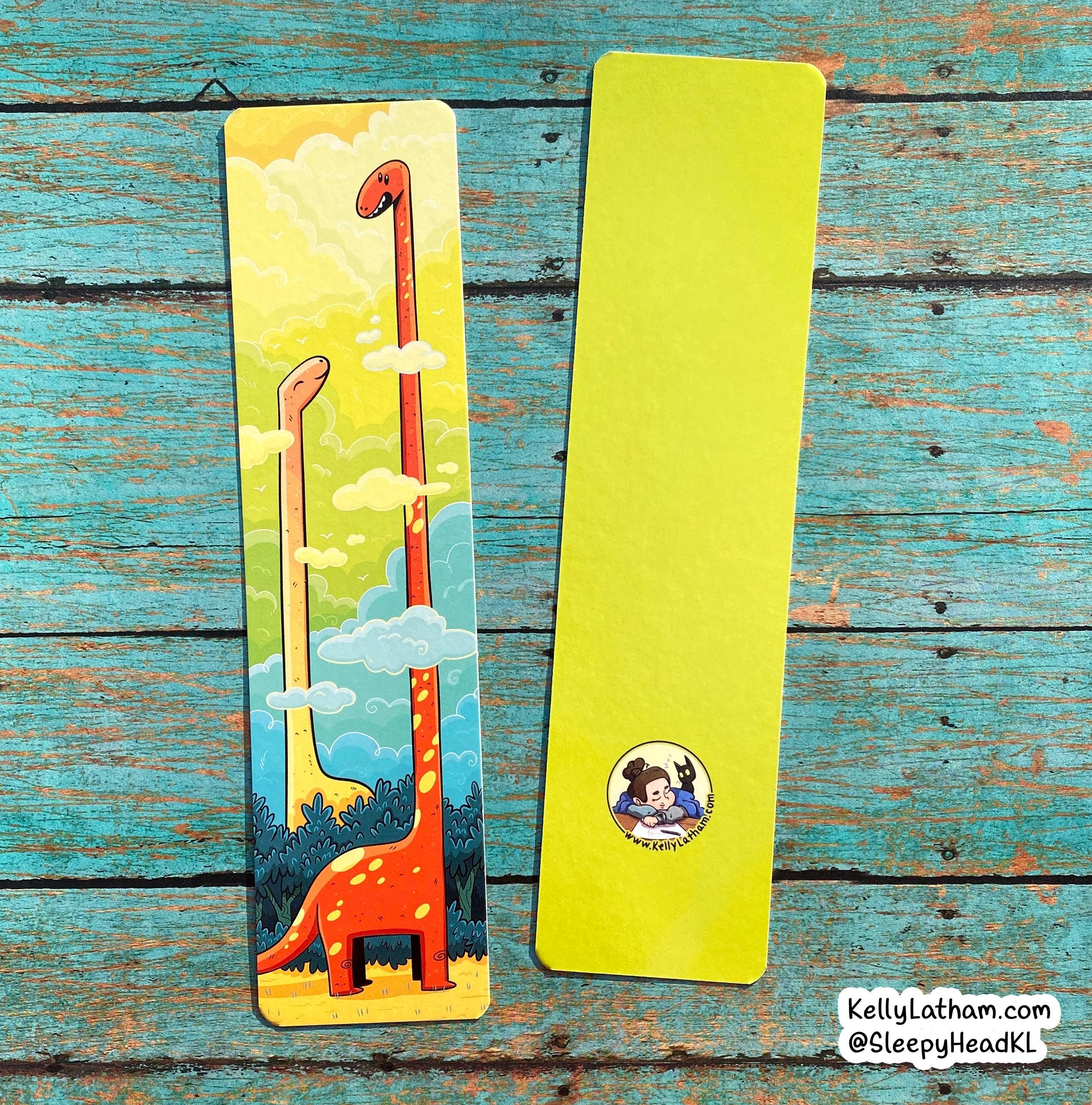 Two slim bookmarks laid flat on a distressed turquoise wood surface. One bookmark is blank bright yellow-green, and the other has a colorful illustration of two long-neck dinosaurs in a sky with clouds.