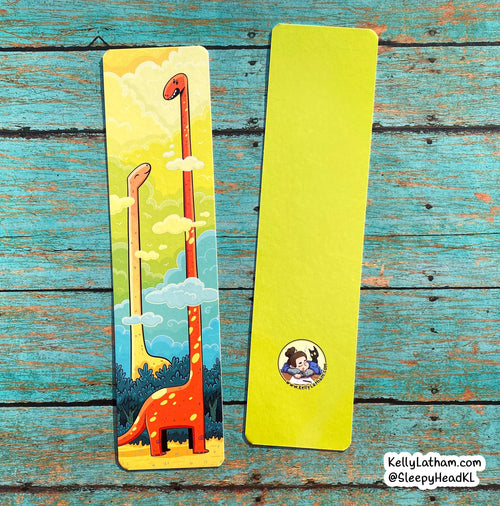 Two slim bookmarks laid flat on a distressed turquoise wood surface. One bookmark is blank bright yellow-green, and the other has a colorful illustration of two long-neck dinosaurs in a sky with clouds.