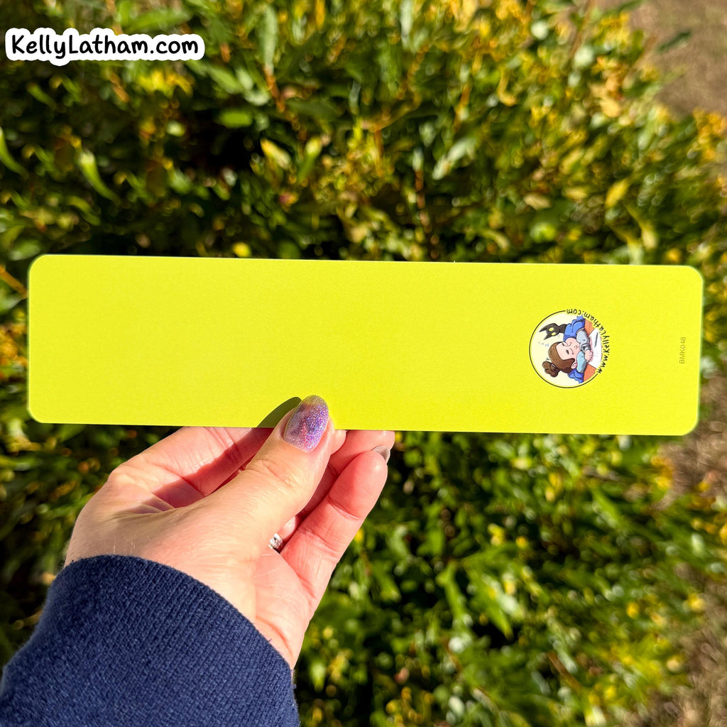 A hand holding a bright yellow-green  bookmark with rounded corners, featuring a small cartoon dog illustration near the top right. The background is a blurred green garden with yellow flowers.