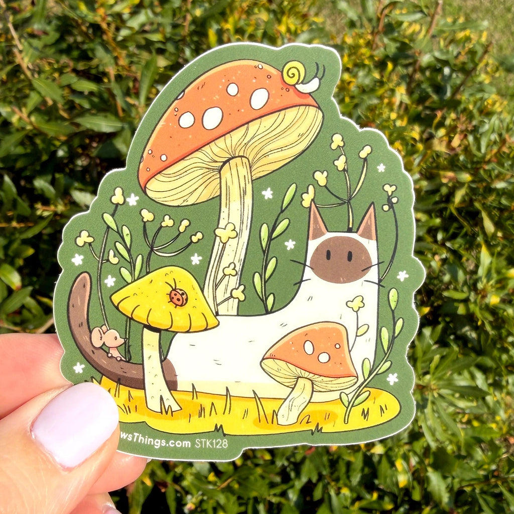A hand holds a die-cut sticker featuring a whimsical forest scene with a white cat, orange mushrooms, green foliage, and a small mouse. The sticker is photographed outdoors against a blurred green hedge background.