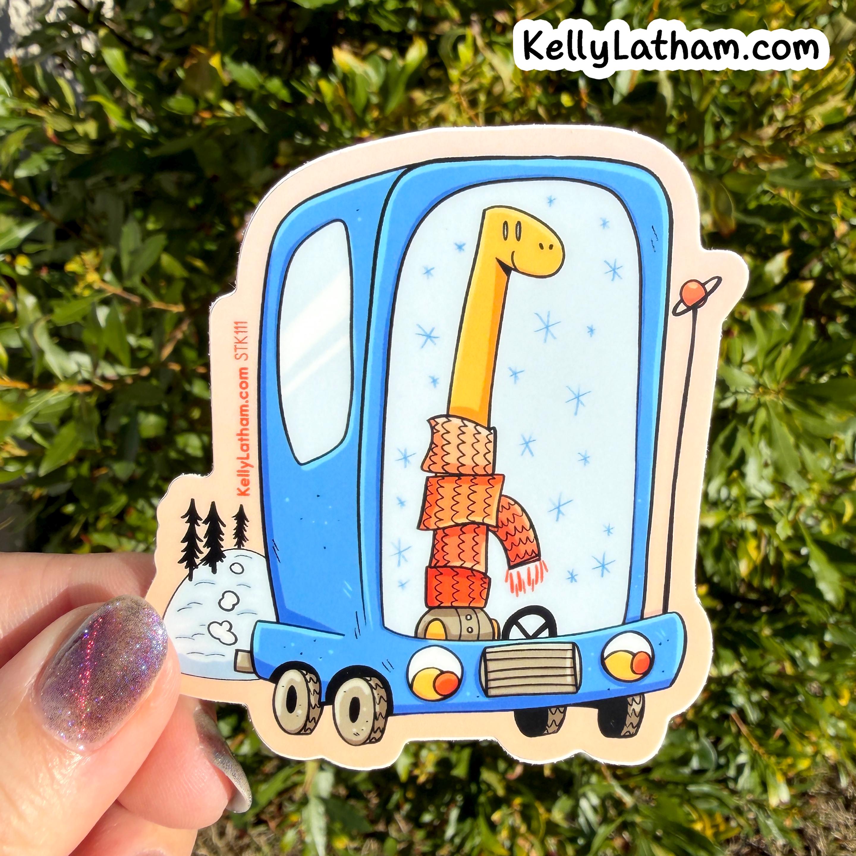 A die-cut sticker held by a hand against a background of green foliage. The sticker features a whimsical illustration of a long-necked yellow dinosaur wearing a red and orange knit sweater inside a blue truck, with snowy trees on the side and the KellyLatham.