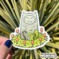 A die-cut sticker featuring a cute gray cat sitting in a grassy field with orange mushrooms, held by a hand against a blurred green plant background.