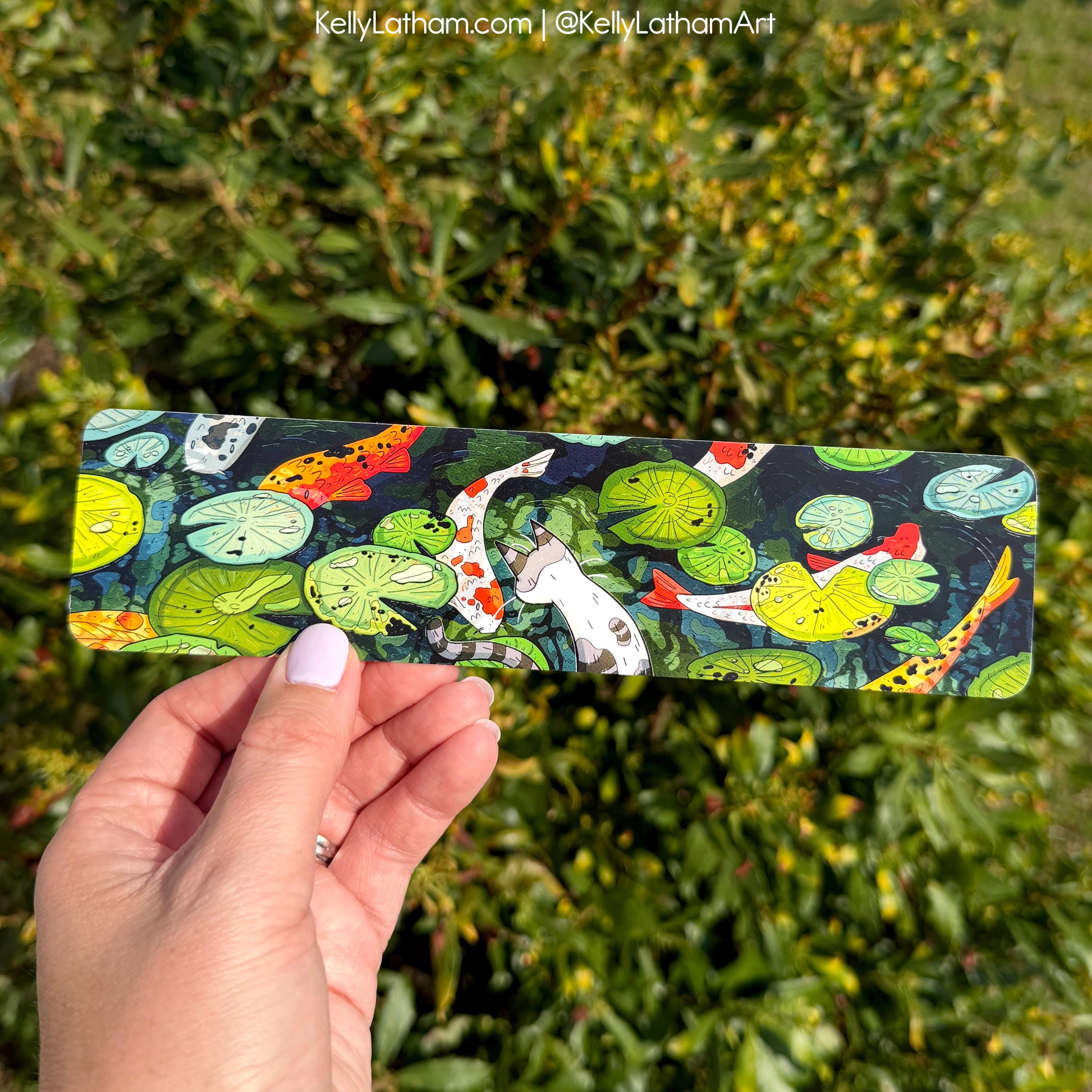 Hand holding a colorful bar of soap with koi fish and lily pads design against a green leafy background.