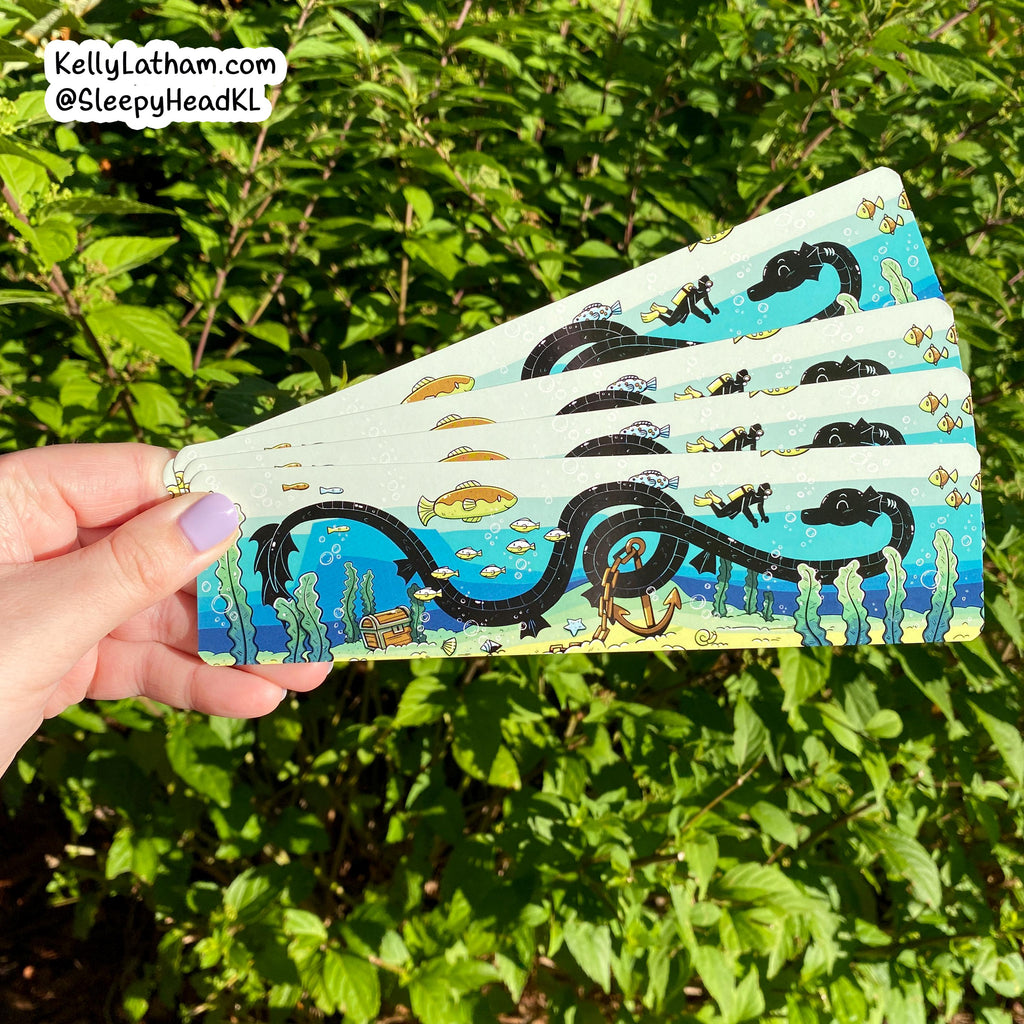 A hand holds a fanned-out set of fabric face masks outdoors in front of green foliage. The masks have a light blue and white ocean-themed print featuring a large black seahorse, fish, divers, and sea plants.