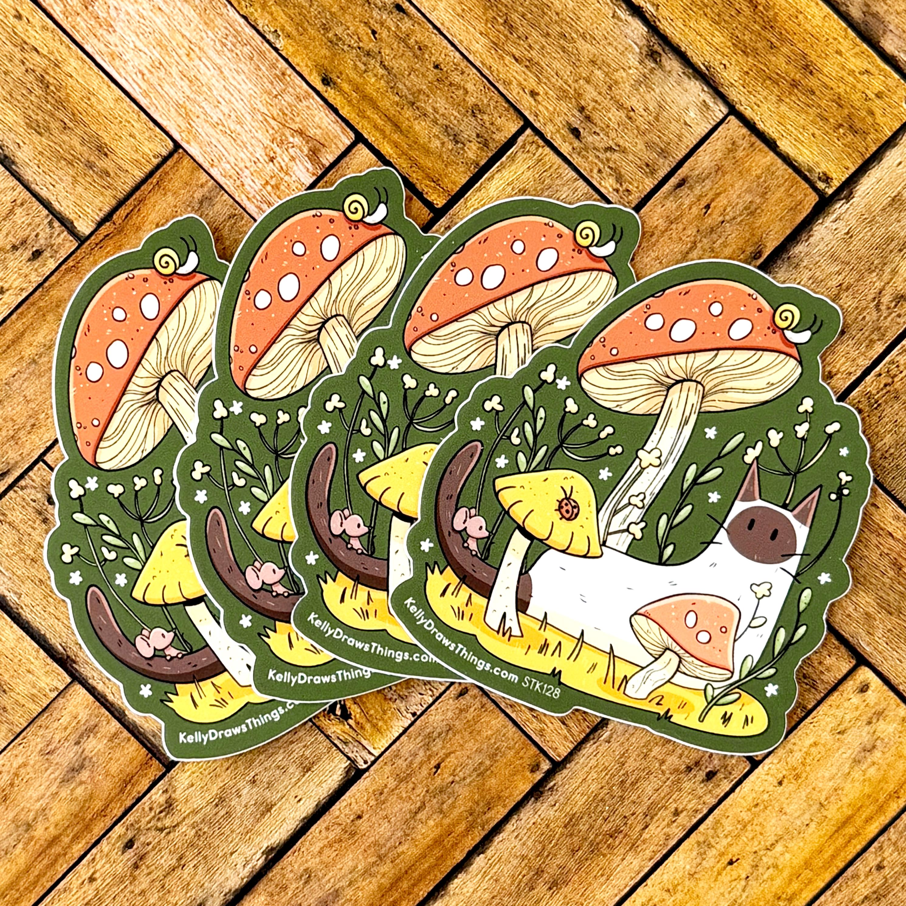 Five die-cut stickers featuring whimsical woodland illustrations of mushrooms, a white cat, and a small dog, arranged in an overlapping fan shape on a rustic wooden plank background. The stickers have green borders and display the brand name 'KellyDrewThings' along with the website 'KellyDrewThings.