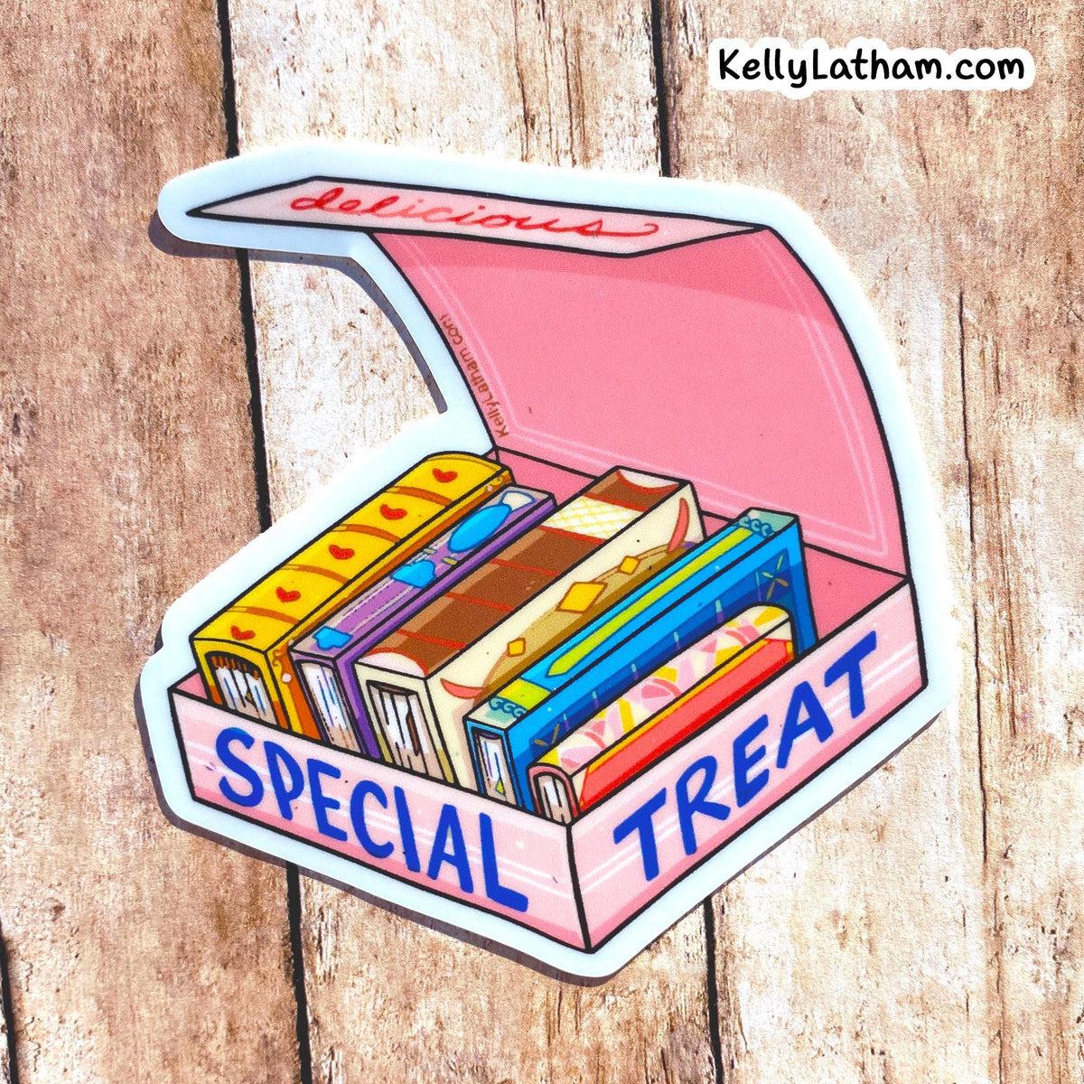 A die-cut sticker depicting an open pink box labeled 'SPECIAL TREAT' filled with colorful illustrated candy bars, placed on a rustic wood background. The brand name 'KellyLatham.