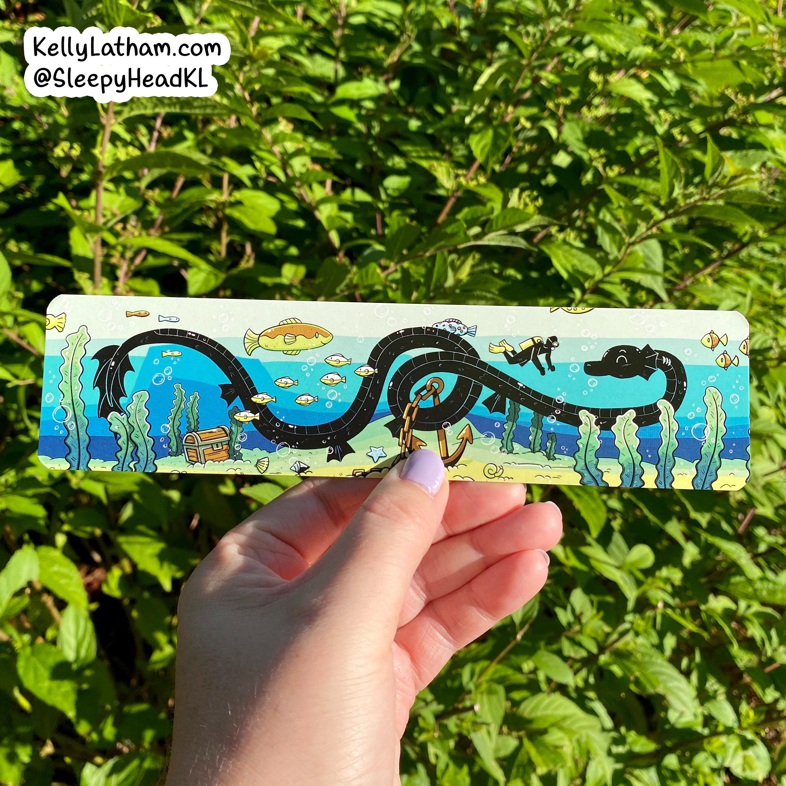 A hand holds a rectangular metal keychain outdoors in front of green foliage. The keychain features a colorful cartoon sea serpent wrapping around a gold anchor, with an underwater scene background.