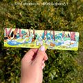 Hand holding a colorful Duck Storytime bookmark featuring illustrated ducks, ducklings, and woodland scenery against a leafy green background.
