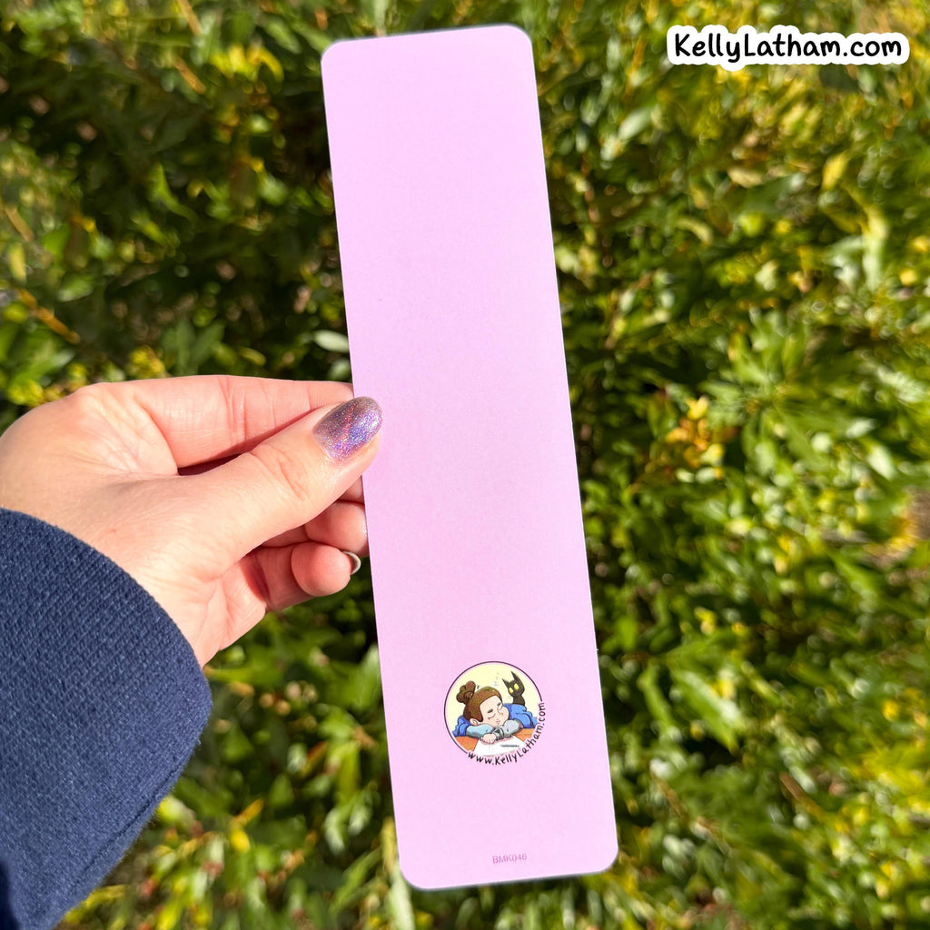 A hand with purple glitter nail polish holds a light pink rectangular bookmark featuring a small circular cartoon illustration of Belle and the Beast from The Princess Bride, set against a background of green foliage.