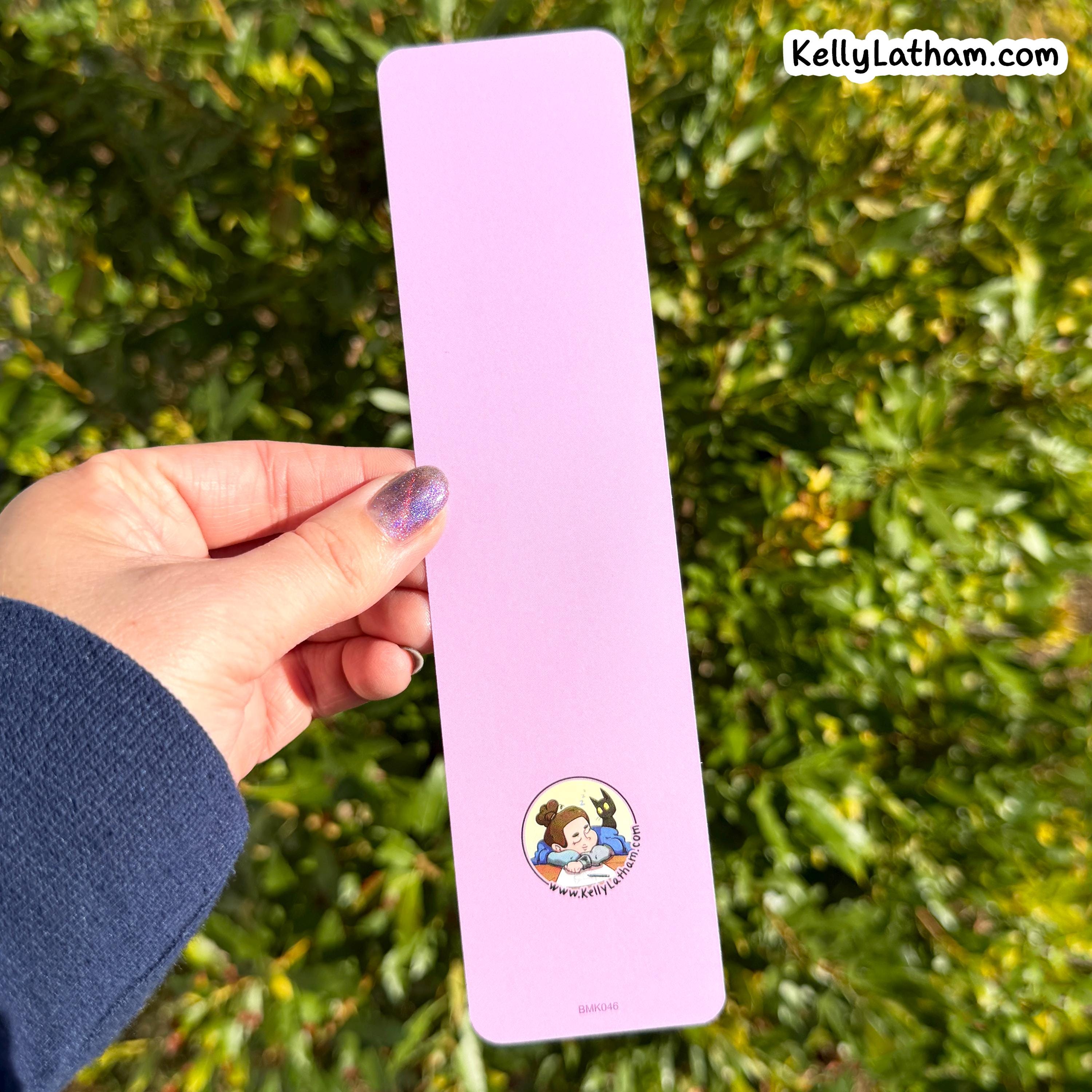 A hand with purple glitter nail polish holds a light pink rectangular bookmark featuring a small circular cartoon illustration of Belle and the Beast from The Princess Bride, set against a background of green foliage.