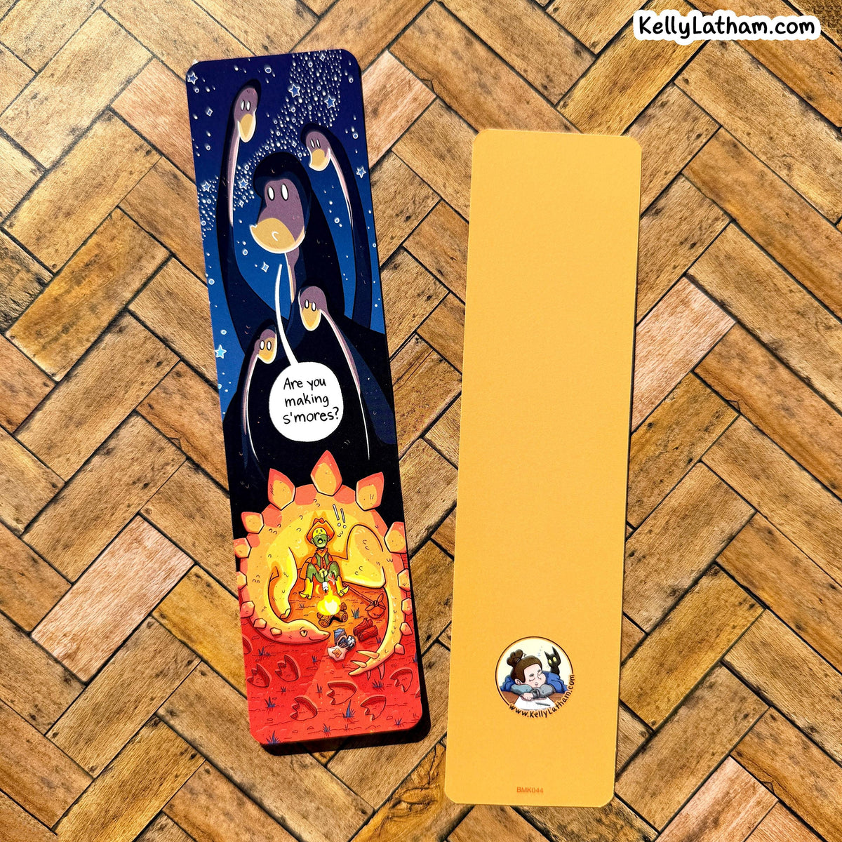 Two tall, narrow bookmarks or card sleeves displayed on a wood herringbone floor. One sleeve has a colorful cartoon illustration with a dark blue upper section, purple bird-like figures, and a red lower section with a small illustrated character.