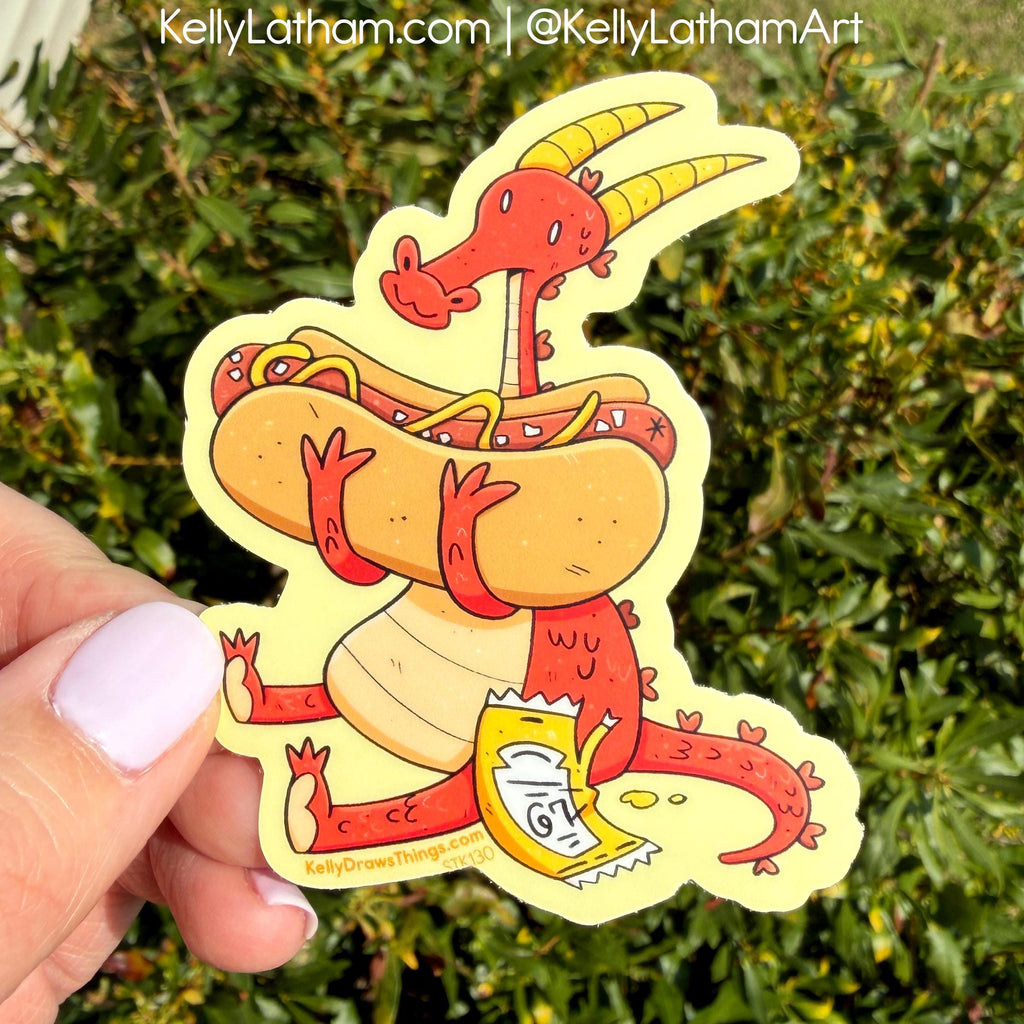 Sticker of a red creature on a hot dog with mustard, held in front of green foliage.