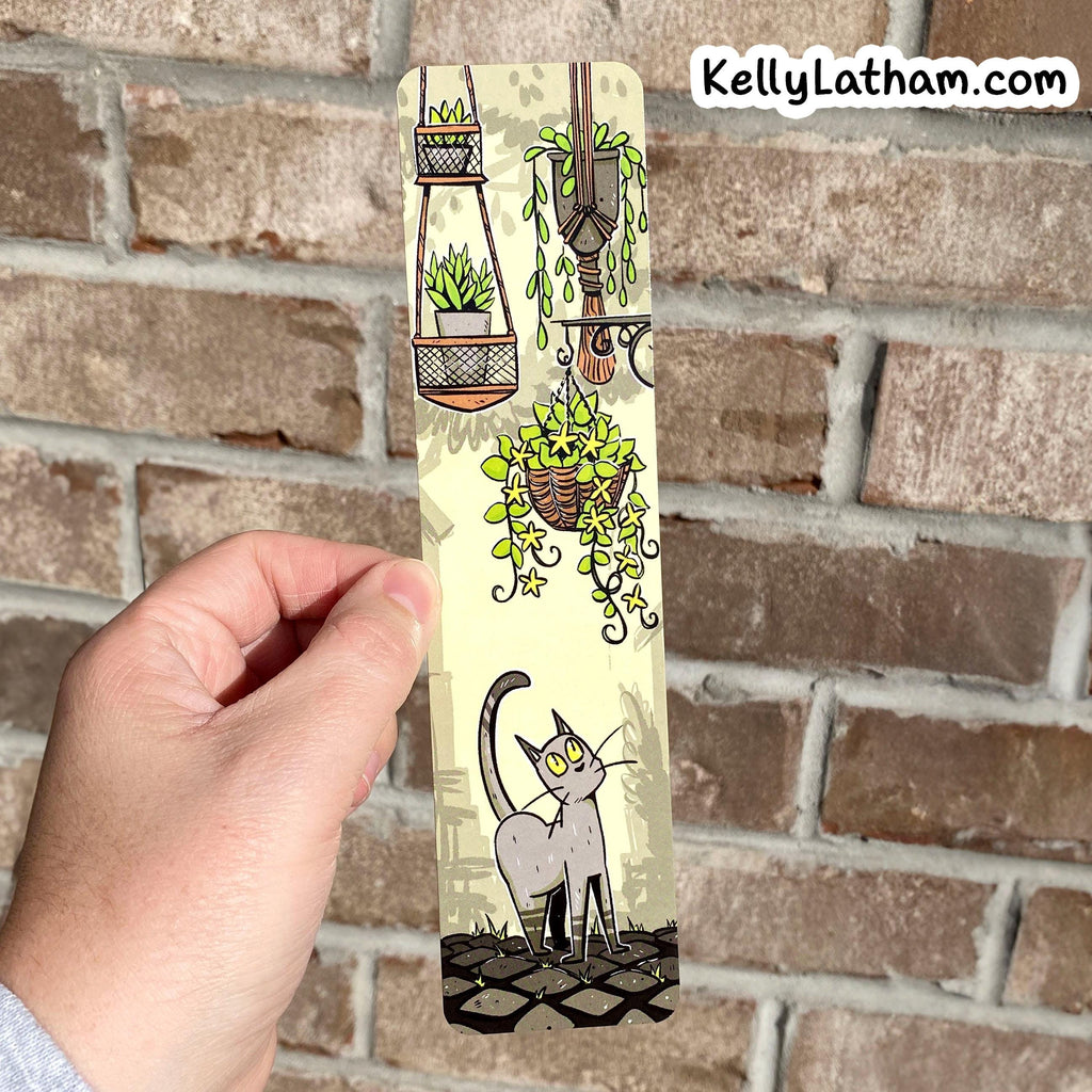 A hand holds a narrow printed card or bookmark featuring an illustration of hanging green plants and a gray cat, against a real brick wall background. The text 'KellyLatham.