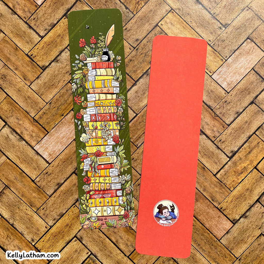 Two decorative paper bookmarks laid flat on a wood herringbone floor. The left bookmark is olive green with an illustrated stack of colorful books, flowers, leaves, and a feather.