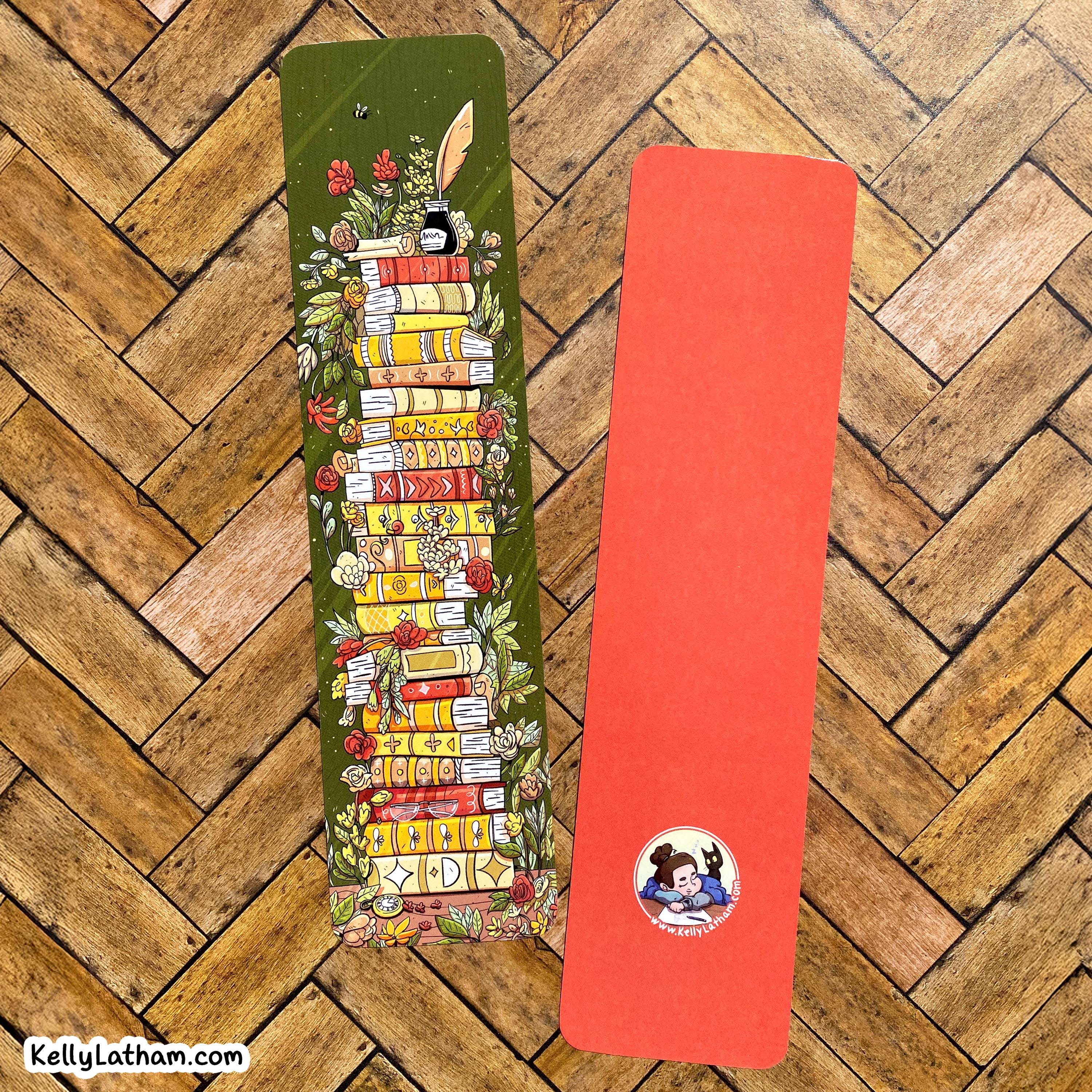 Two decorative paper bookmarks laid flat on a wood herringbone floor. The left bookmark is olive green with an illustrated stack of colorful books, flowers, leaves, and a feather.