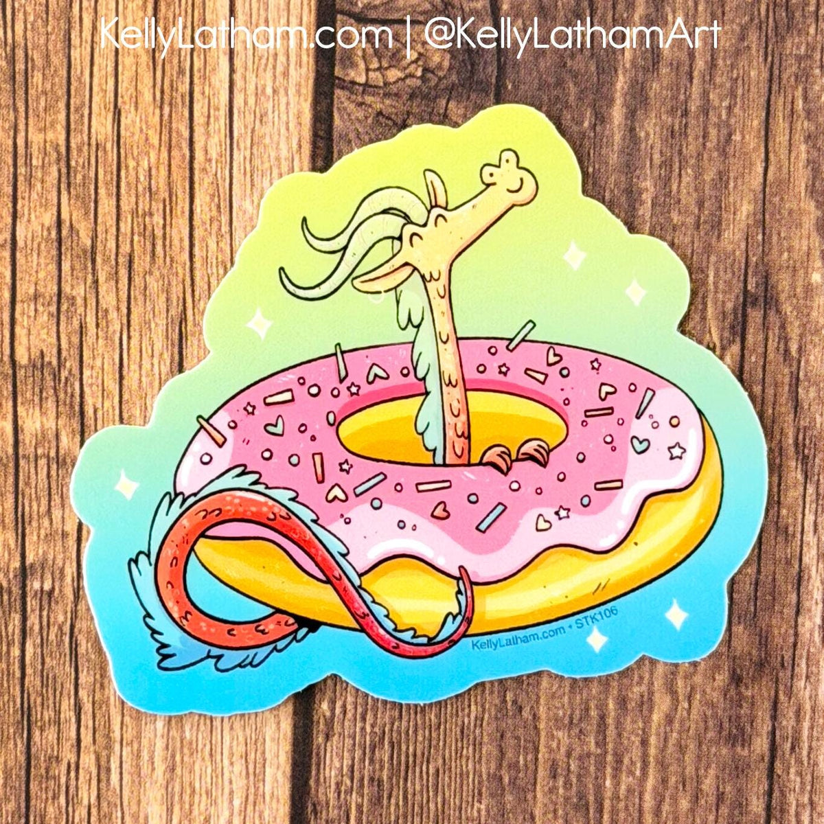 A die-cut sticker featuring a whimsical illustration of a pink frosted donut with sprinkles and a long-necked dragon rising from the center, placed on a wooden surface. The sticker has a blue border and watermark text at the top and bottom.