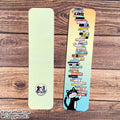 Two decorative bookmarks laid flat on a wood surface. One bookmark is plain pale yellow-green with a small circular character sticker near the bottom.