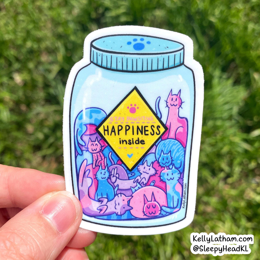 A hand holds a die-cut sticker shaped like a light blue cat jar, featuring a yellow diamond label that reads 'HAPPINESS inside' and colorful cartoon cats in pink, blue, and purple. The background is a blurred green outdoor setting.