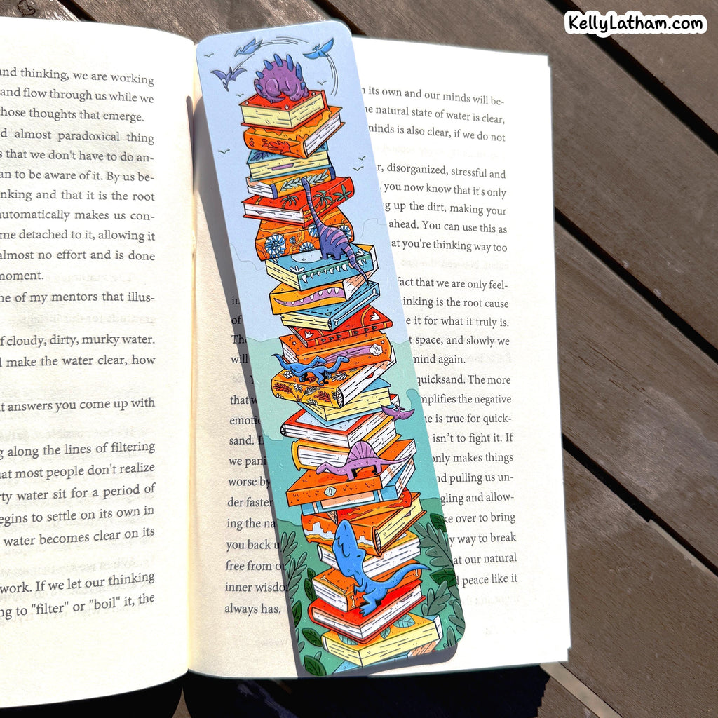 A decorative paper bookmark shaped like a stack of colorful books with cartoon birds and a blue sky, placed across an open book on a dark wooden surface.