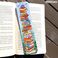 A decorative paper bookmark shaped like a stack of colorful books with cartoon birds and a blue sky, placed across an open book on a dark wooden surface.