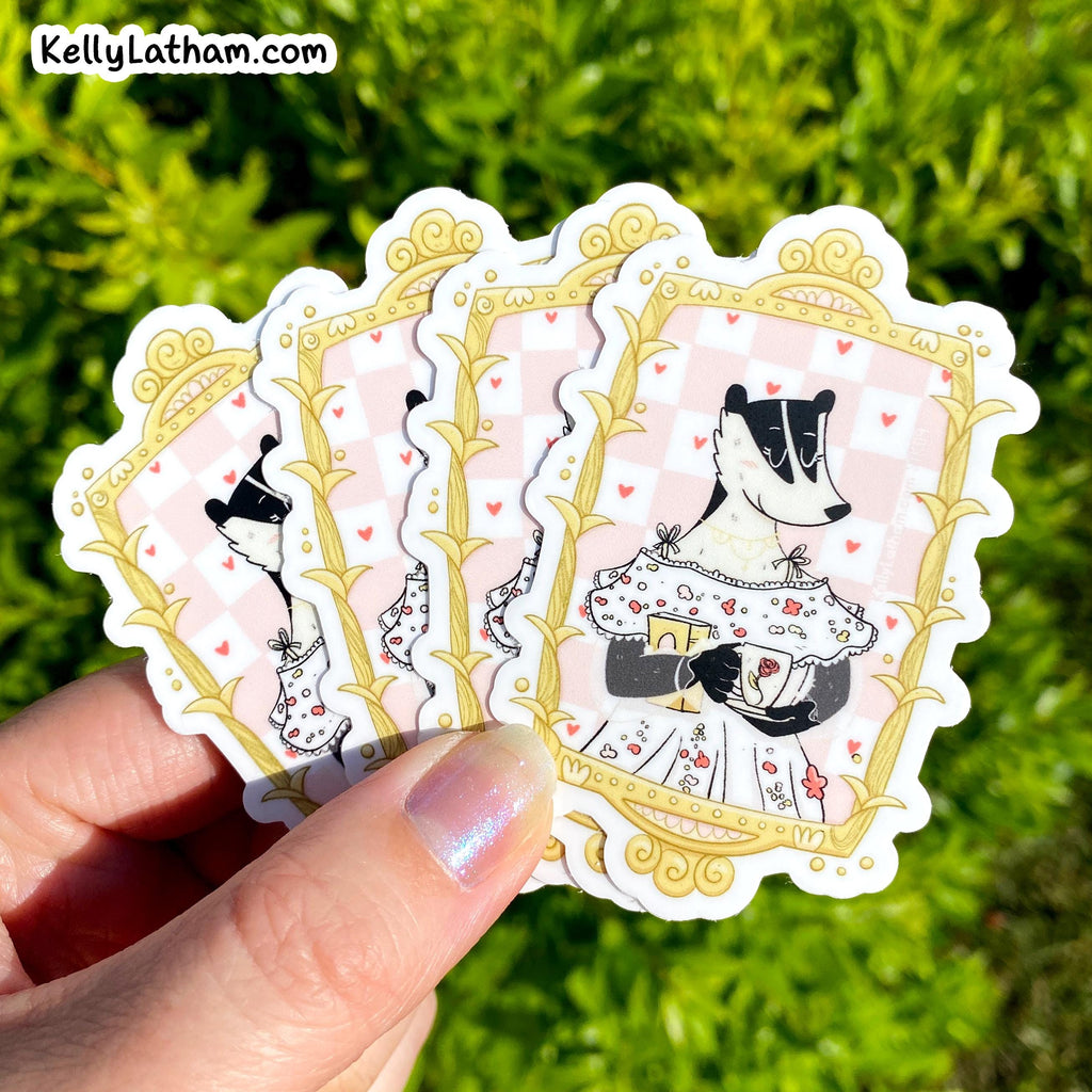 A hand holds a fan of four die-cut stickers outdoors against a blurred green grass background. The stickers feature a black-and-white badger in a white dress with red hearts, framed by ornate gold borders on a pale pink check background.