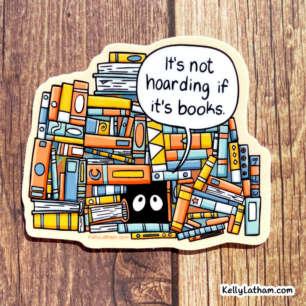 A die-cut sticker featuring an illustrated pile of colorful books with a speech bubble that reads, 'It's not hoarding if it's books.' The sticker is photographed on a wood surface.