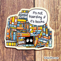 A die-cut sticker featuring an illustrated pile of colorful books with a speech bubble that reads, 'It's not hoarding if it's books.' The sticker is photographed on a wood surface.