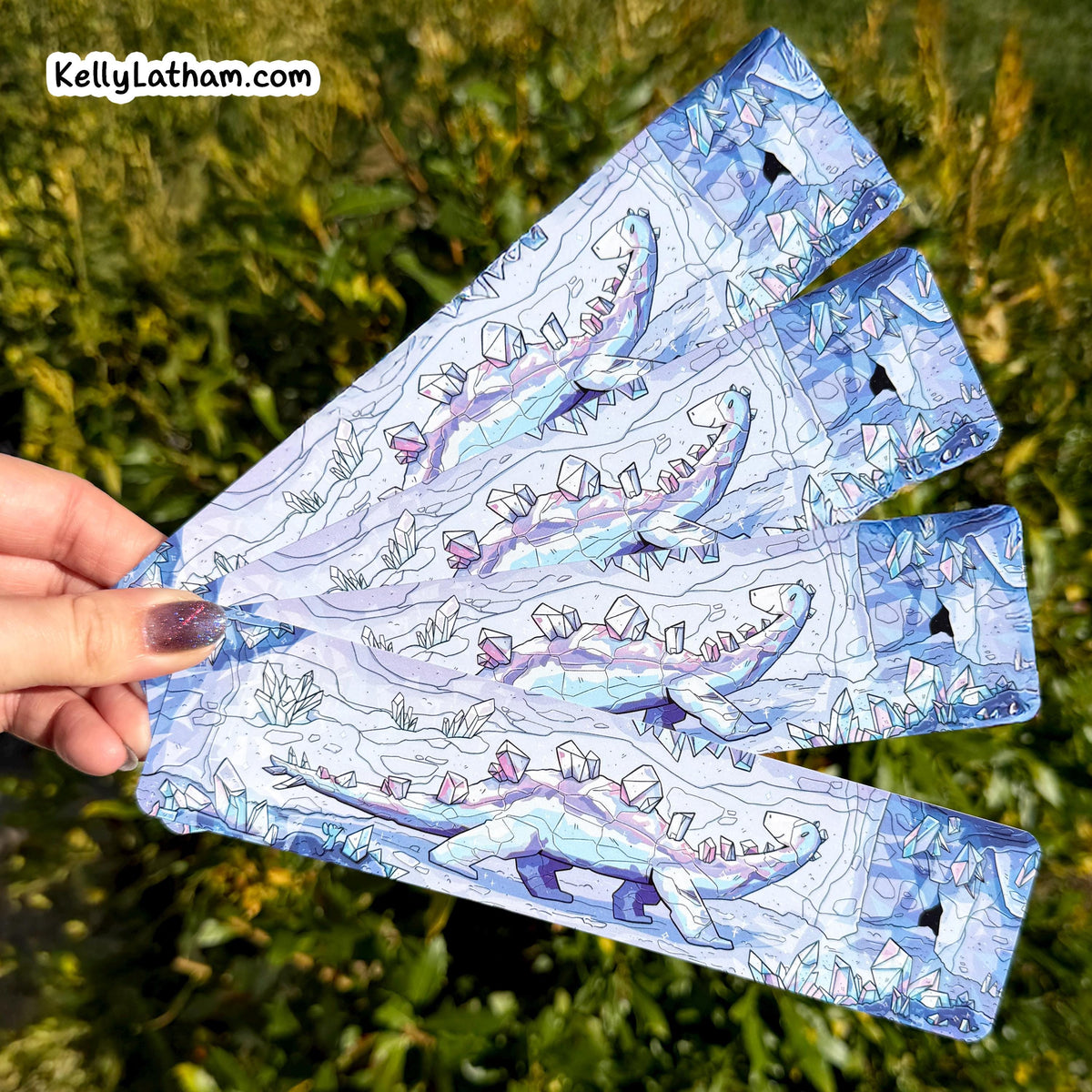 A hand holds a fan of four rectangular printed cards or sleeves outdoors, with green foliage in the background. The cards have a blue and white illustrated pattern with line-art style graphics and small black triangular cutouts near the edges.