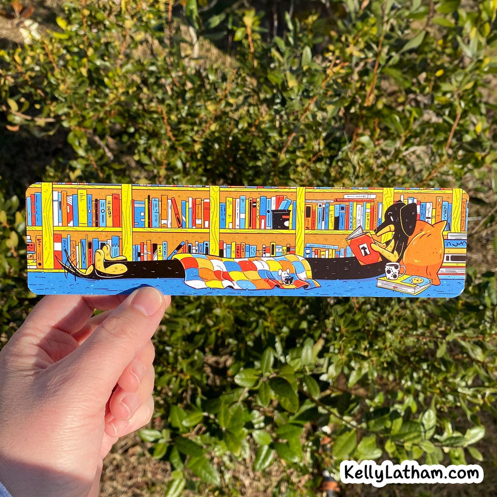 A hand holds a long rectangular printed card or book cover featuring a colorful illustrated library scene with bookshelves, books, a cat, and a dog. The background is real outdoor greenery, and a small 'KellyLatham.